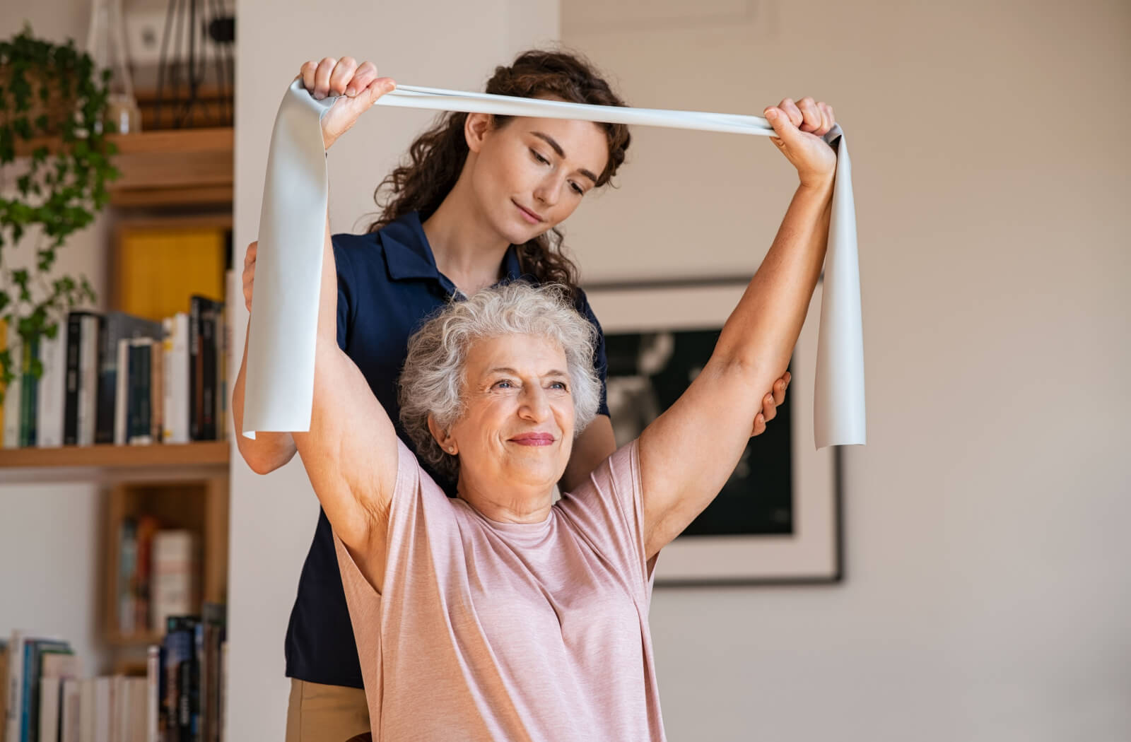 An older adult in senior living holding a resistance band overhead to improve joint health with their caregiver's support.