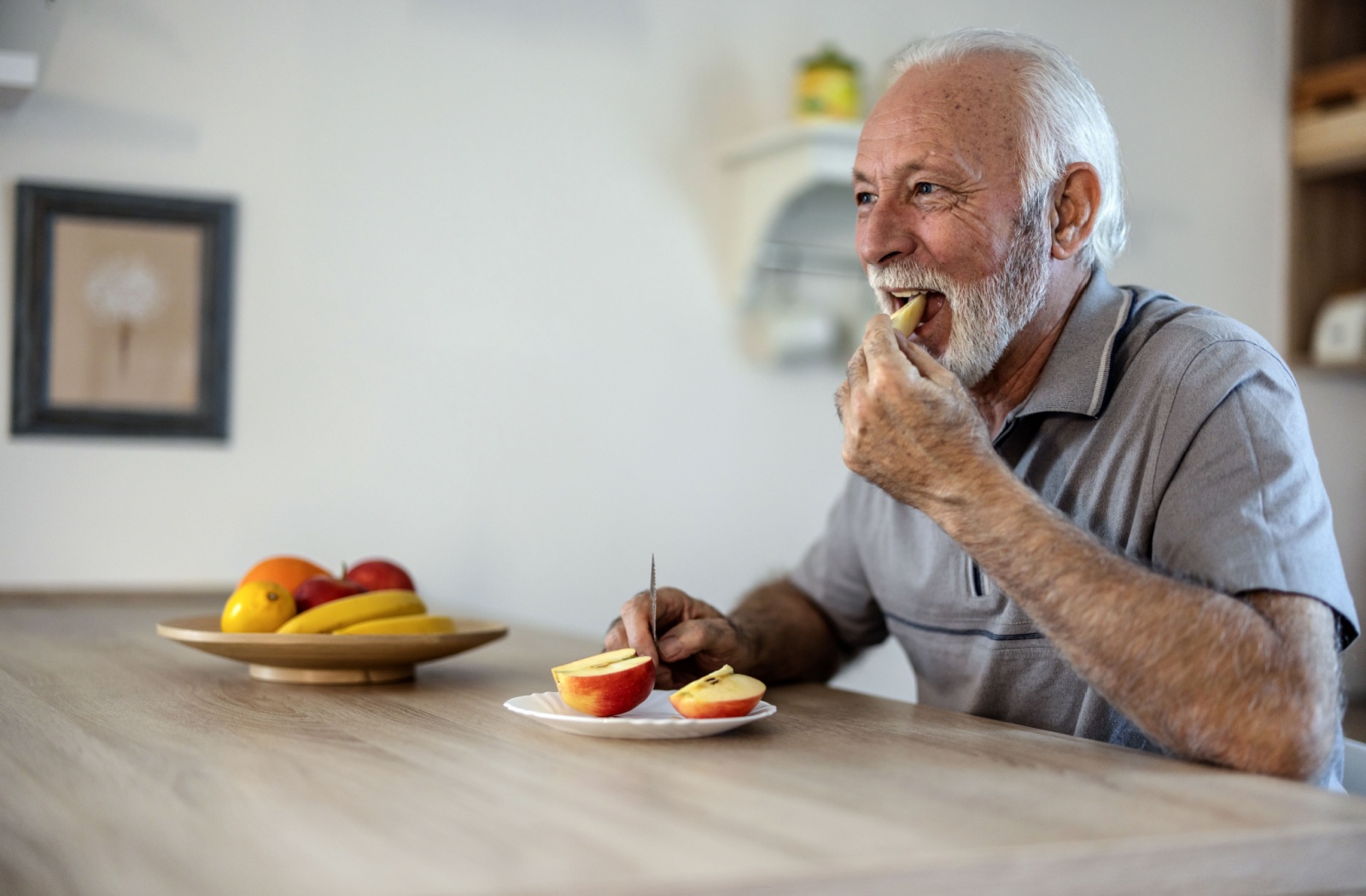 An older adult sits in a bright kitchen next to a plate of vibrant fruit, eating a nutritious, healthy apple.