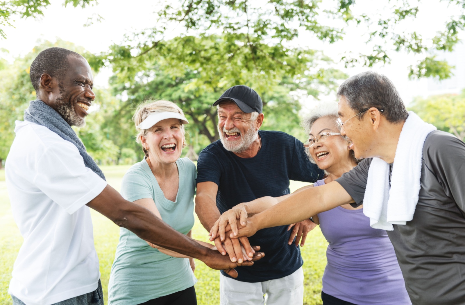 A group of smiling seniors in activewear stand outdoors with their hands stacked together, showing teamwork and social connection.