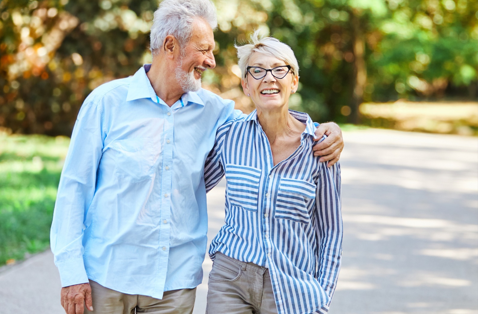 An older adult and their spouse smile while walking down a path in a park with arms around one another