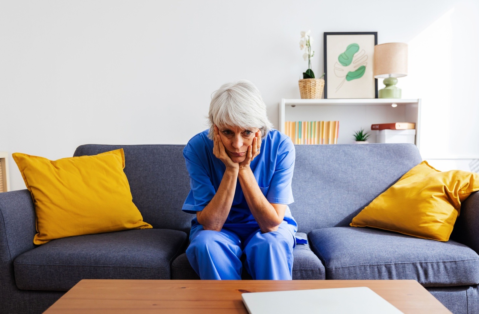 An adult caregiver in blue scrubs looking defeated while sitting on a grey sofa with bright yellow pillows
